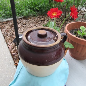 May include: A brown and white glazed ceramic crock with a lid. The crock is sitting on a blue and white patterned fabric. There are red flowers in the background.
