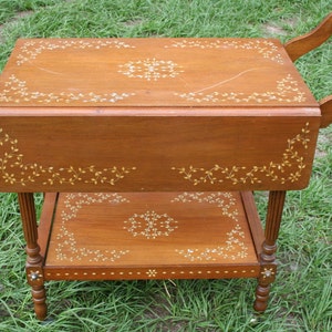 May include: A wooden serving cart with two tiers. The top tier has a drop-leaf design and both tiers feature intricate floral patterns inlaid with mother-of-pearl.