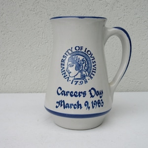 May include: Cream-colored ceramic pitcher with a blue rim and handle. The pitcher features a blue emblem of the University of Louisville and the text "Careers Day March 9, 1963". A vintage collectible item.