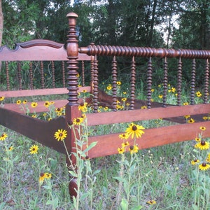 May include: A vintage wooden bed frame with a unique, intricate design featuring a series of turned wooden spindles. The bed frame is surrounded by a field of yellow wildflowers.