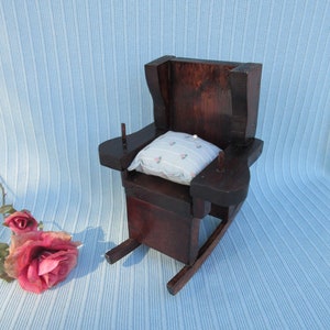 May include: A small wooden rocking chair with a white and pink floral cushion. The chair is dark brown and has a simple design.