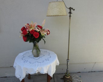 May include: An antique floor lamp with a textured lampshade stands next to a small round table. The table is covered with a white lace tablecloth and holds a vase of red roses and pink lilies. The lamp has a decorative metal base.