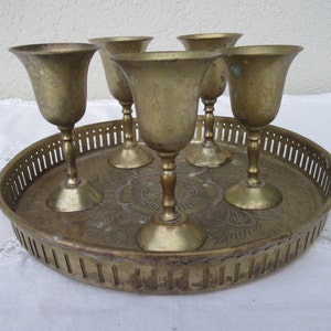 May include: A set of five gold-toned metal shot glasses on a matching tray with an ornate design. The tray has a decorative, openwork border.