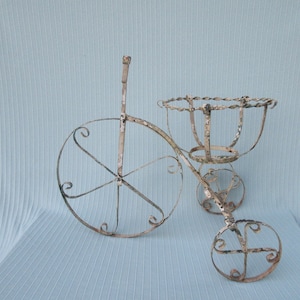 May include: White metal plant stand shaped like a tricycle with three wheels and a basket on top. The stand is distressed and has a rustic look.