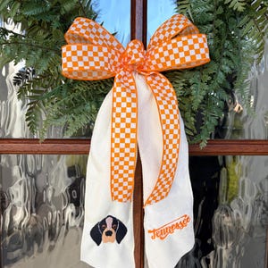 May include: A decorative wreath with a large orange and white checkered bow. The wreath includes a white fabric sash with an embroidered dog and the word "Tennessee" in orange script. Green foliage frames the wreath.