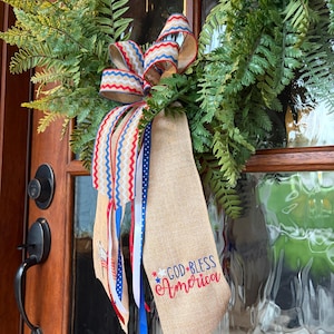 May include: A green fern wreath with a patriotic bow and ribbons. The bow features red, white, and blue chevron patterns. A burlap ribbon is embroidered with "GOD BLESS America" in red and blue.