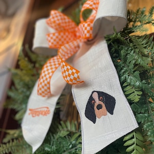 May include: A decorative bow featuring a hound dog design. The bow has a white textured ribbon with an embroidered hound dog face and the word "Tennessee". The bow also has orange and white checkered ribbon accents, set against a backdrop of green foliage.