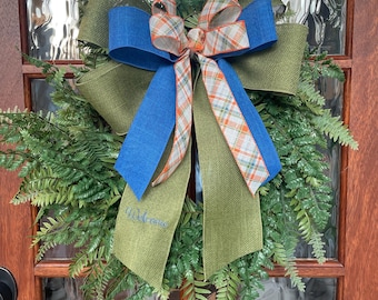 May include: A fern wreath with a large bow. The bow has blue, green, and plaid ribbons. The green ribbon has the word "Welcome" embroidered on it. The wreath is hanging on a wooden door with glass panels.
