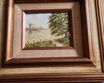 May include: A framed landscape painting depicting a rural scene with a farmhouse, windmill, and a large tree. The painting is set within a double-layered wooden frame with a light beige inner layer and a darker brown outer layer.