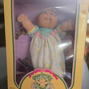 May include: A Cabbage Patch Kids Preemie doll in a white and blue floral dress with a pink bow. The doll is in a clear plastic box with a yellow and purple background. The box has the text "1985 Cabbage Patch Kids Preemie!" and "I'm unique. I'm adoptable!" The box also has the March of Dimes logo.