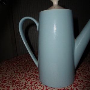 May include: A light blue ceramic coffee pot with a curved handle and a spout. The lid is white and beige. The pot is on a red and white patterned tablecloth.