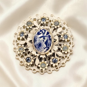 May include: A vintage white and blue floral brooch. The brooch features a central oval porcelain piece with a blue floral design. It is surrounded by a white filigree frame with blue and clear crystal accents. The brooch is a classic piece of jewelry.