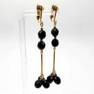 May include: Clip-on earrings with a gold-tone metal design. Each earring features a gold clip, a small gold bead, two faceted black beads, a gold bar, and a cluster of black beads at the bottom. The earrings are displayed on a clear stand.