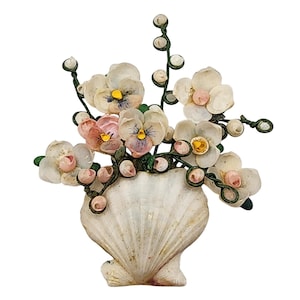 May include: A decorative seashell vase filled with delicate, handcrafted flowers. The flowers are primarily white with yellow and purple centers, and some have pink accents. The vase is a natural, off-white color, and the stems are green.
