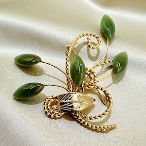 May include: A gold-toned brooch featuring a floral design with five green, oval-shaped jade stones. The brooch has a textured, twisted gold wire design and a detailed gold leaf-shaped element at the base.