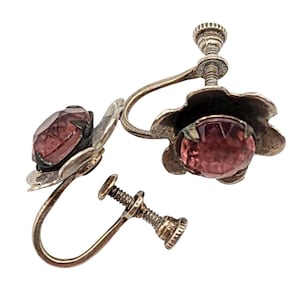 May include: A pair of silver-toned screw-back earrings featuring a flower-shaped metal setting with a central round, faceted, dark pink gemstone. The metal has a darkened, aged patina finish, and the screw mechanism is clearly visible on each piece.