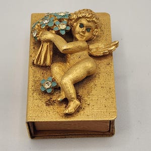 May include: A rectangular gold-toned metal matchbox cover featuring a three-dimensional cherub figure holding a bouquet of light blue enameled flowers with clear rhinestone centers. The cherub has wings and a textured gold finish throughout.