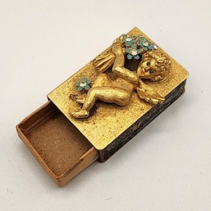 May include: A vintage gold-tone matchbox with an embossed cherub design holding flowers. The cherub is adorned with turquoise and clear rhinestones. The matchbox is open, revealing the interior.