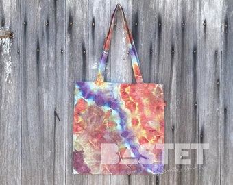 May include: A tie-dye tote bag with a colourful design in shades of orange, yellow, blue, and purple. The bag has a white fabric label with the text "BASTET by emilie b" printed on it.