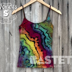 Pode incluir: Um top de renda tie-dye com um padrão de redemoinho arco-íris em tons de vermelho, laranja, amarelo, verde, azul e preto. O top está pendurado em um cabide de madeira contra um fundo de madeira. O texto "BASTET by michelle" é visível na parte inferior da imagem.