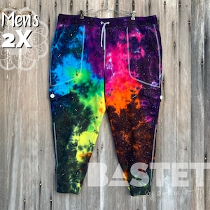 Op de afbeelding: Regenboog tie-dye sweatpants met zwart-witte galaxy print en witte stiksels. De sweatpants hebben twee grote cargozakken aan de zijkanten. De sweatpants zijn gelabeld "Men's 2X" en "BASTET by Emily D."