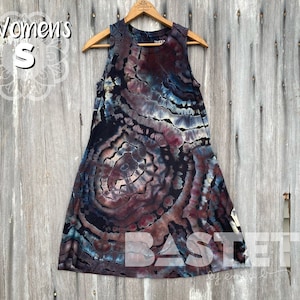 Pode incluir: Um vestido sem mangas de tie-dye com um padrão circular em tons de azul, marrom e preto. O vestido está pendurado em um cabide de madeira sobre um fundo de madeira. O texto "Women's S" é visível no canto superior esquerdo da imagem. O texto "BASTET by emily b" é visível no canto inferior direito da imagem.