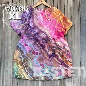 Peut inclure: Tunique tie-dye XL aux couleurs vives, notamment rose, violet, orange et bleu. La tunique a des manches courtes et une taille froncée. Le texte "Women's XL" est visible en haut à gauche. La tunique est suspendue à un cintre en bois.