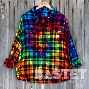 Könnte beinhalten: Langärmliges, kariertes Hemd in Regenbogenfarben mit Knopfleiste. Das Hemd zeigt eine lebendige Mischung aus Farben, darunter Rot, Orange, Gelb, Grün, Blau und Lila. Es hängt an einem Holzbügel vor einem Holz Hintergrund.