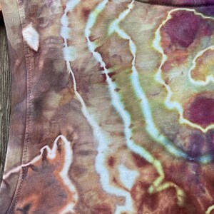 May include: A close-up of a brown, green, and purple tie-dye tank top. The fabric has a swirling pattern with light and dark shades of brown, green, and purple.