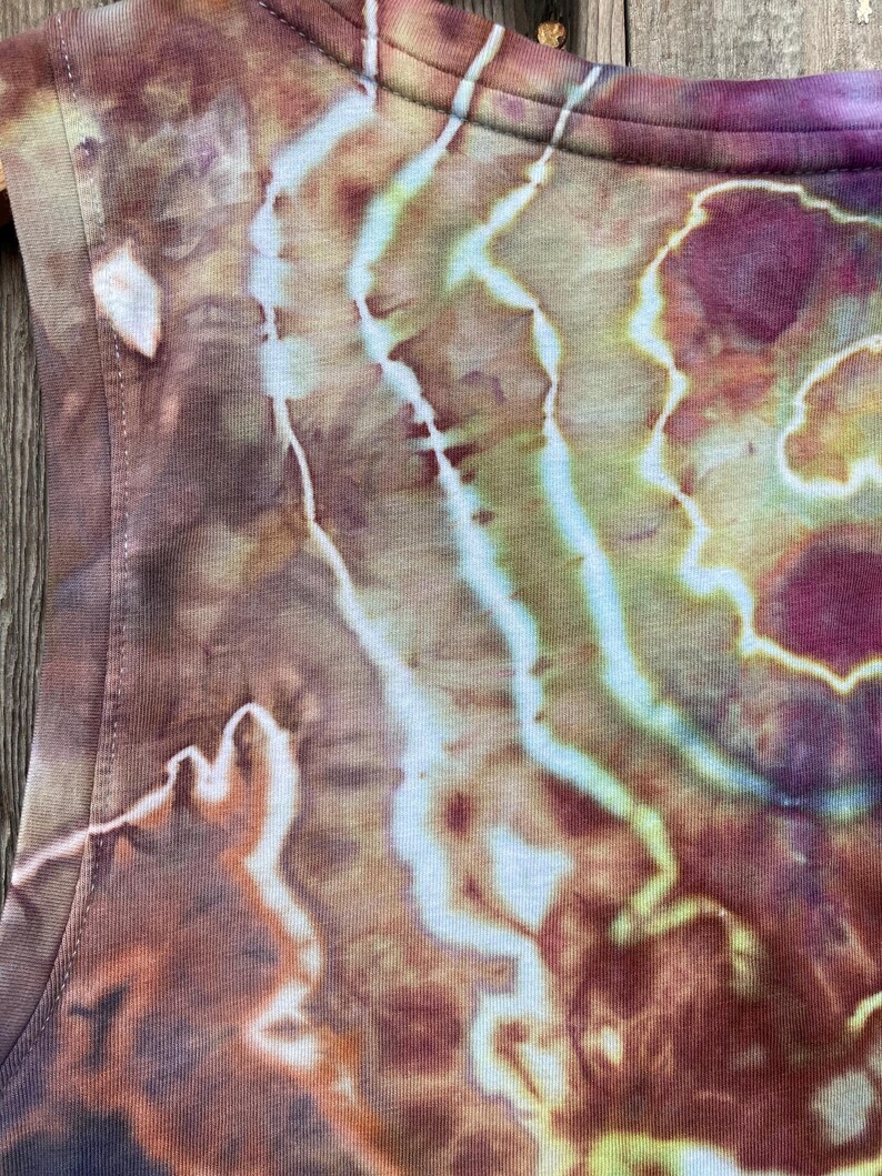 May include: A close-up of a brown, green, and purple tie-dye tank top. The fabric has a swirling pattern with light and dark shades of brown, green, and purple.