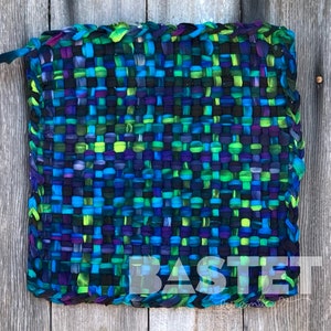 May include: A woven rug with a blue, green, and purple color scheme. The rug is made of fabric strips and has a fringed edge. The rug is labeled "BASTET by Emilia D."