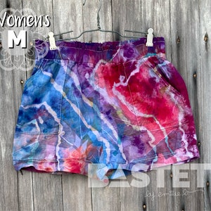 May include: Tie-dye shorts in shades of blue, purple, and red, with a white pattern. The shorts have pockets and an elasticated waistband. The text "Womens M" is visible on the top left. The shorts are hanging on a metal hanger.