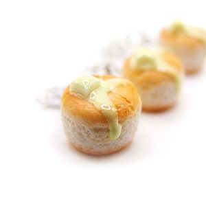 以下が含まれることがあります： 白いアイシングと薄い茶色の皮をした、ミニチュアの丸い焼き菓子が3つ。焼き菓子は白い表面に置かれており、ポリマークレイでできているように見える。