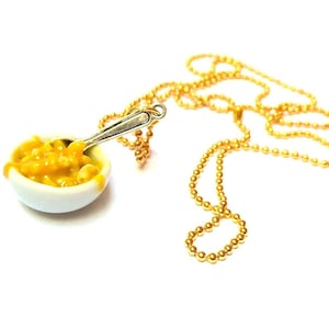May include: A gold chain necklace with a small white bowl filled with yellow macaroni and cheese and a silver spoon.