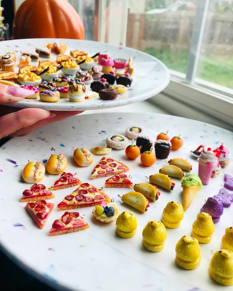 K&ouml;nnte beinhalten: Eine Sammlung von Miniatur-Lebensmittel-Anh&auml;ngern, darunter Pizzast&uuml;cke, Tacos, Hotdogs, Eiswaffeln, Cupcakes, Donuts und vieles mehr. Die Anh&auml;nger sind aus Polymer Clay gefertigt und haben ein realistisches Aussehen.