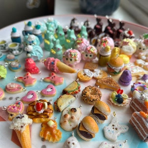 K&ouml;nnte beinhalten: Eine Sammlung bunter Polymer Clay Charms mit verschiedenen Lebensmittel- und Tiermotiven. Die Charms werden auf einem wei&szlig;en Teller pr&auml;sentiert und beinhalten Artikel wie Eiswaffeln, Hamburger, Donuts und Hasen.