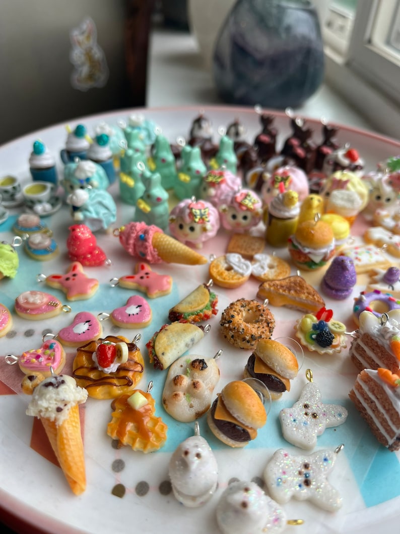 K&ouml;nnte beinhalten: Eine Sammlung bunter Polymer Clay Charms mit verschiedenen Lebensmittel- und Tiermotiven. Die Charms werden auf einem wei&szlig;en Teller pr&auml;sentiert und beinhalten Artikel wie Eiswaffeln, Hamburger, Donuts und Hasen.