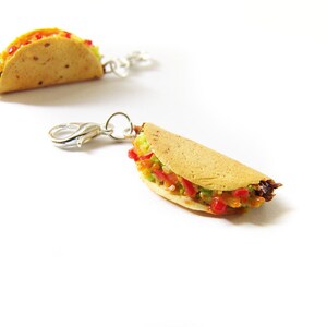 K&ouml;nnte beinhalten: Zwei Miniatur-Taco-Anh&auml;nger mit silbernen Verschl&uuml;ssen. Die Tacos sind mit Salat, Tomate und Fleisch gef&uuml;llt.