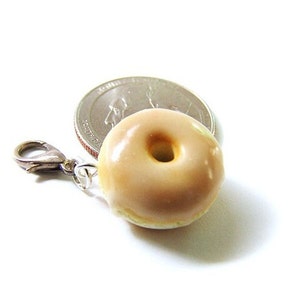 May include: A small, glazed donut charm with a silver clasp. The donut is light brown and has a white glaze. The charm is about the size of a quarter.