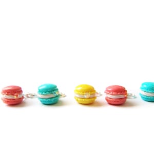 Könnte beinhalten: Fünf bunte Macaron-Anhänger in Rot, Türkis, Gelb, Koralle und Türkis. Jeder Anhänger hat eine weiße Füllung und einen silberfarbenen Ring. Die Macarons sind in einer Reihe vor weißem Hintergrund angeordnet.