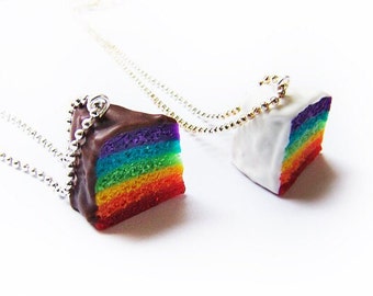 May include: Two rainbow cake slice necklaces. One slice is topped with chocolate frosting and the other with white frosting. Both necklaces have silver chains.