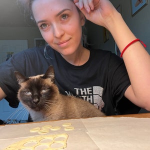 K&ouml;nnte beinhalten: Eine Frau mit einer Katze auf ihrem Scho&szlig;, beide schauen in die Kamera. Die Frau tr&auml;gt ein schwarzes T-Shirt mit dem Text "The North Face" darauf. Die Katze ist ein Siam mit blauen Augen. Vor ihnen liegt ein St&uuml;ck Teig mit ausgeschnittenen Kreisen auf einer wei&szlig;en Oberfl&auml;che.