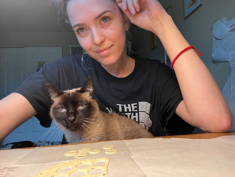 K&ouml;nnte beinhalten: Eine Frau mit einer Katze auf ihrem Scho&szlig;, beide schauen in die Kamera. Die Frau tr&auml;gt ein schwarzes T-Shirt mit dem Text "The North Face" darauf. Die Katze ist ein Siam mit blauen Augen. Vor ihnen liegt ein St&uuml;ck Teig mit ausgeschnittenen Kreisen auf einer wei&szlig;en Oberfl&auml;che.