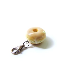 May include: A small, glazed donut charm with a silver clip. The donut is light brown with a white glaze.