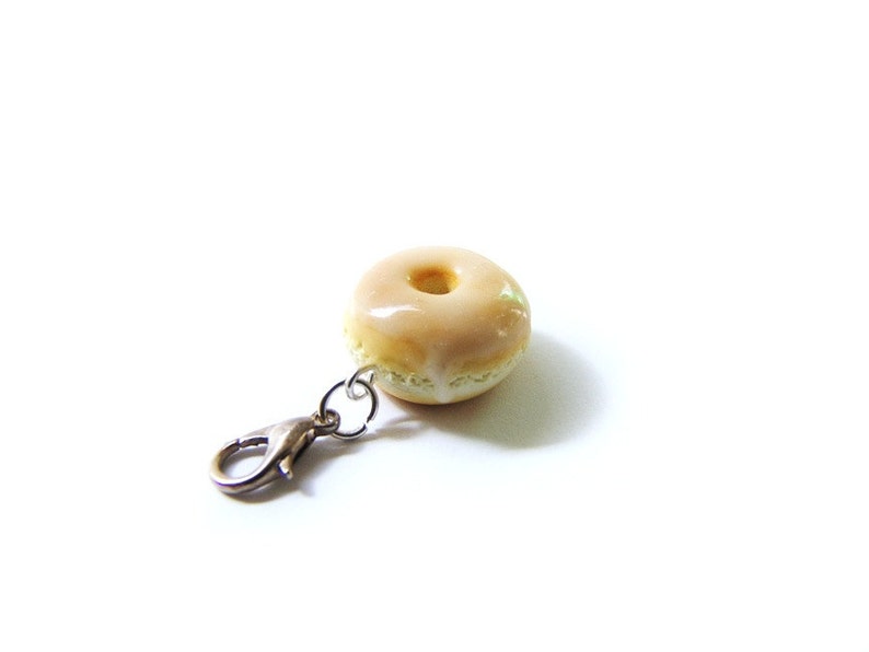 May include: A small, glazed donut charm with a silver clip. The donut is light brown with a white glaze.