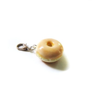May include: A glazed donut charm with a silver metal clasp. The donut is light brown with a white center.