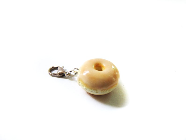 May include: A glazed donut charm with a silver metal clasp. The donut is light brown with a white center.