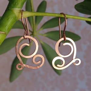 May include: A pair of rose gold earrings with a delicate, swirling heart design. The earrings are made of wire and have a hammered texture.