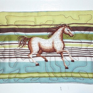 May include: A brown and white horse embroidered on a striped fabric cushion with green, brown, and blue stripes. The cushion is made of a soft, quilted fabric.