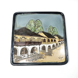 May include: Square ceramic decorative plate with a black border. The plate features a detailed painting of a building with arched walkways, brown roofs, and green trees against a blue sky. The artist's signature, "Viga", is visible.